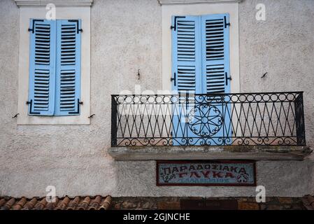 Ländliche Hausfassade mit hellblauen Holzfensterläden und einem Balkon mit einem handgefertigten geschmiedeten Eisengeländer an einer verwitterten weißgetünchten Wand in Galaxidi GR Stockfoto