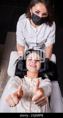 Schöne Brünette Frau, die Daumen hoch und lächelnd, auf dem Daybed liegend und mit Zustimmung Geste, während Arzt Kosmetikerin in sterilen Handschuhen hinter ihr sitzen. Konzept der Spa-Behandlung und Hautpflege. Stockfoto
