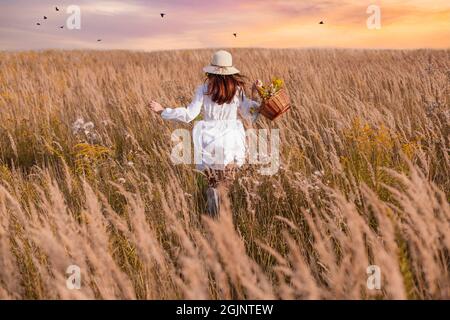 Eine schöne Frau mit einem Korb mit Kräutern läuft in einem Blumenfeld. Herbalistisches Konzept, Glück. Stockfoto