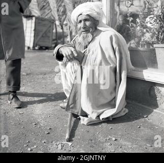 Der historische, lokale Afghane der 1950er Jahre, mit Bart, Kopfschmuck und Decke über ihm sitzend, hockend in einer Straße in der Nähe von Kabul, Afghanistan. Stockfoto