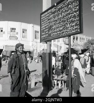 1950, historisch, Afghanistan, Kabul, draußen in einer Straße, ein afghanischer Mann, der neben einem Schild oder einer Tafel steht, sowohl auf Englisch als auch auf Arabisch, für das Munzil Bagh Hotel. In der Schrift steht, dass das Hotel offers......comfortable Unterkünfte ... alle Einrichtungen für Sommer und Winter, afghanische und europäische Küche, Swimmingpool und Tennisplatz. Stockfoto