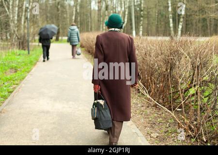 Eine alte Frau geht die Straße hinunter. Ein Rentner im Mantel geht durch den Park. Verheiratet von hinten in Herbstkleidung. Meine Großmutter in Russland ist es Stockfoto