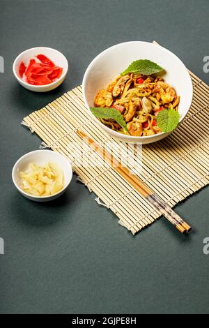 WOK-Nudeln mit Meeresfrüchten und Gemüse. In Öl gebratene Muscheln, Garnelen und Tintenfische. Köstliches asiatisches Mittagessen in einer weißen Schüssel auf einer Bambusmatte. Braten Sie Noo unter Rühren an Stockfoto