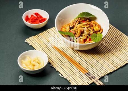 WOK-Nudeln mit Meeresfrüchten und Gemüse. In Öl gebratene Muscheln, Garnelen und Tintenfische. Köstliches asiatisches Mittagessen in einer weißen Schüssel auf einer Bambusmatte. Braten Sie Noo unter Rühren an Stockfoto