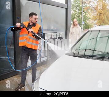 Junger Mann, der im Freien an der Autowaschstation Auto wascht, trägt eine orangefarbene Weste. Hübscher Arbeiter, der Auto putzt, mit Hochdruckwasser. Stockfoto