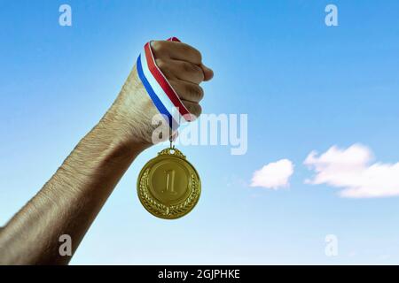 Goldmedaille in der Siegerhand. Erster Platz auf klarem blauen Himmel Hintergrund, Sport Champion Athlet Sieg Konzept Stockfoto