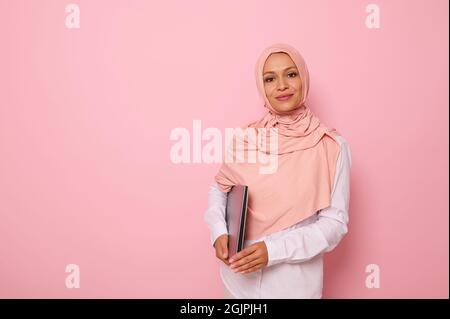 Isoliertes, selbstbewusstes Geschäftsporträt auf farbigem Hintergrund einer arabischen muslimischen hübschen Frau mit überdecktem Kopf in rosa Hijab, trägt ein weißes Hemd und sieht aus Stockfoto