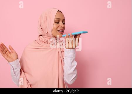 Muslim Arab wunderschöne schöne Frau mit verdecktem Kopf in rosa Hijab Aufnahme einer Sprachnachricht auf ihrem Smartphone. Isoliertes Porträt auf rosa pastellfarbenem Bac Stockfoto