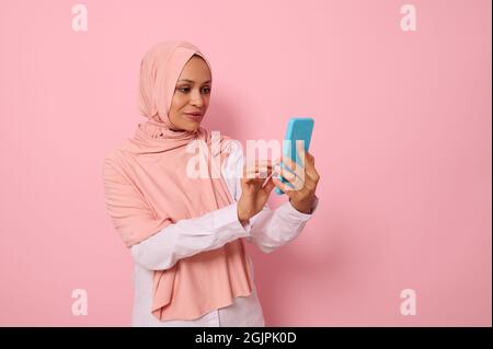 Isoliertes Porträt einer selbstbewussten arabisch-muslimischen reifen hübschen Frau in streng religiösem Outfit und verdecktem Kopf in rosa Hijab SMS-Nachricht auf einem Handy Stockfoto