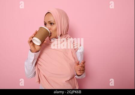 Schöne arabische muslimische Frau, die einen Hijab trägt und heißes Getränk, Tee oder Kaffee aus Einweg-Pappbecher trinkt, seitlich stehend, drei Quart Stockfoto