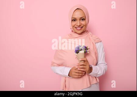 Isoliertes Porträt auf farbigem Hintergrund einer schönen muslimischen arabischen Frau in rosa Hijab, mit einem Strauß Wildblumen in lila Farbtönen, eingewickelt Stockfoto