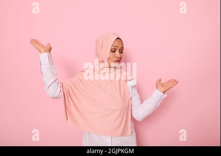 Hüftlanges Porträt einer wunderschönen, geheimnisvollen muslimischen Frau, die einen Kopftuch-Hijab trägt und etwas auf ihren Handflächen zeigt, isoliert auf einem farbigen rosa Rückenband Stockfoto