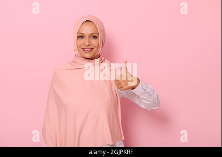 Isoliertes Porträt auf rosa Hintergrund mit Kopffreiche einer entzückenden und charmanten arabischen Frau in Hijab zeigt einen Daumen nach oben und lächelt mit schönen t Stockfoto