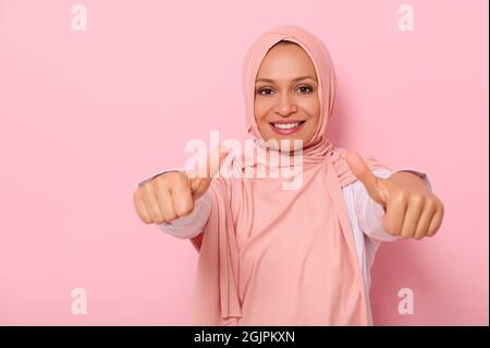 Arabisch-muslimische freundliche und charmante Frau in rosa Hijab zeigt Daumen nach oben auf die Kamera, lächelt mit toothy Lächeln, isoliert auf farbigem Hintergrund mit s Stockfoto