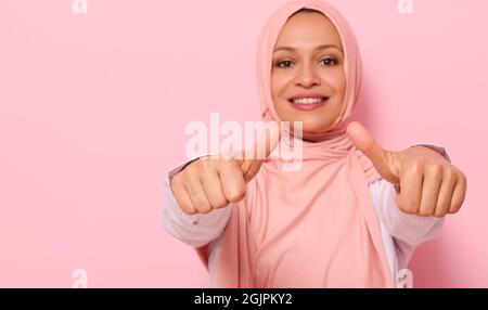 Weicher Fokus auf die Hände eines verschwommenen Lächelns mit toothy Lächeln arabische muslimische Frau in rosa Hijab, zeigt Daumen nach oben auf die Kamera, isoliert über farbigen Bac Stockfoto