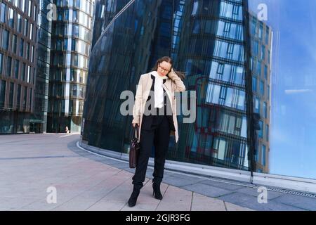 Eine Geschäftsfrau aus dem Kaukasus in einem Mantel und Anzug mit nachdenklicher Optik steht in einer Brille in der Nähe des Geschäftszentrums. Das Konzept der Lösung komplexer Geschäftsprobleme Stockfoto