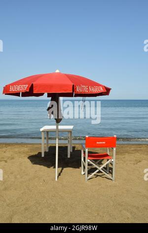 Im Sommer steht ein Rettungsschwimmer mit rotem Regenschirm und Regiestuhl am Sandstrand der toskanischen Maremma, Marina di Castagneto Carducci Stockfoto