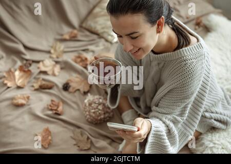 Eine Frau benutzt das Telefon und hält eine Tasse Tee, während sie im Bett in einem gemütlichen Strickpullover sitzt, verschwommener Hintergrund mit Herbstblättern. Stockfoto