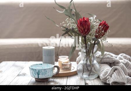 Tasse, Kerzen, Vase mit protea-Blumen und einem gestrickten Element im Raum auf verschwommenem Hintergrund. Stockfoto