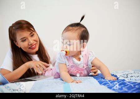 Lächelnde junge Frau, die mit ihrer kleinen Tochter auf dem Bett liegt und versucht, am Laptop zu arbeiten Stockfoto