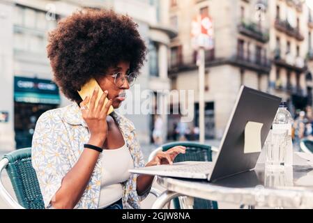 afrikanische Frau sitzt auf einer Barterrasse und telefoniert. Sie benutzt einen Laptop. Stockfoto