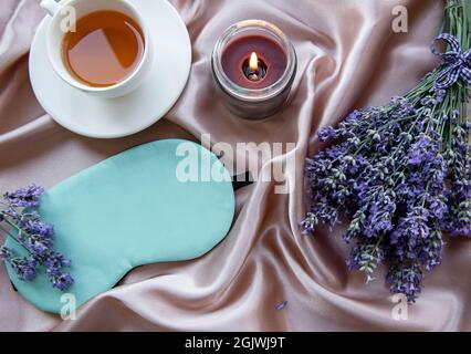 Konzept für gesunden Schlaf. Lavendelbouquet, Schlafmaske und Kerze auf Seidenhintergrund Stockfoto