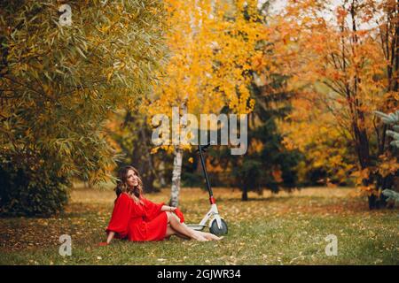 Junge hübsche Frau mit Elektroroller in rotem Kleid sitzen auf Gras im Herbst Stadtpark. Stockfoto