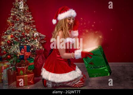 Nettes Mädchen als Weihnachtsmann gekleidet, Blick in einer Geschenktüte mit einem Glanz und Sterne, hinter dem Weihnachtsbaum und Geschenke. Stockfoto