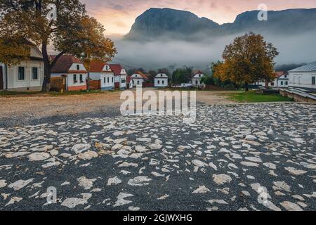 Toller Blick auf die ländliche Straße mit traditionellen weiß getünchten Häusern in Reihe bei Sonnenaufgang. Torocko Dorf und Piatra Secuiului Berg im Hintergrund bei Sonnenaufgang, Stockfoto