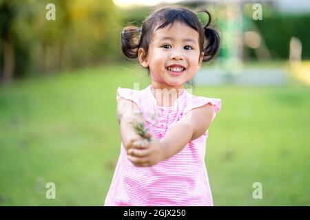 Ein hübsches thailändisches Kind mit pinkem Kleid, das lächelt und einen Blumenstrauß im grünen Feld hält Stockfoto
