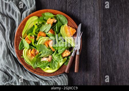 Lachs Spinat Avocado und grüner Pfeffer Salat auf einem braunen rustikalen Teller mit Gabel und Messer auf einem Mooreichentisch, mediterrane gesunde Ernährung, flaches Lay, Fr. Stockfoto