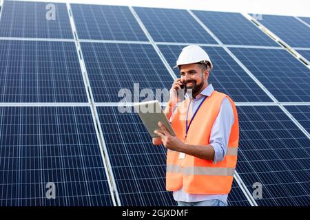 Der junge Elektriker in Weste und Helm hält ein Tablet und telefoniert. Es steht vor dem Hintergrund von Solarzellen Stockfoto