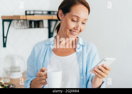 Junge attraktive lächelnde Frau mit einer Tasse Kaffee kommuniziert Nachrichten am Telefon. Stockfoto
