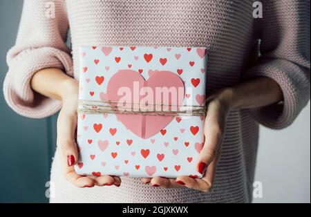 Eingewickeltes Geschenk zum Valentinstag mit einem großen rosa Herz in weiblichen Händen dekoriert. Stockfoto