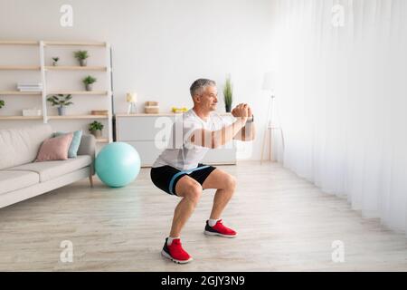 Morgendliches Training. Sportlich reifer Mann tut Kniebeugen, Training im Wohnzimmer Interieur, kopieren Raum Stockfoto