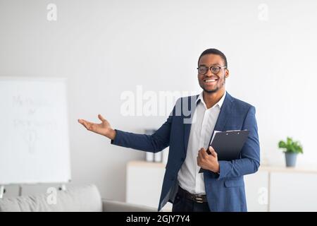 Freundlicher junger afroamerikanischer Psychologe mit Clipboard, der etwas mit seiner Hand in der psychiatrischen Klinik zeigt Stockfoto