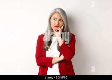 Unruhige asiatische Geschäftsfrau in roten Blazer und Lippen, Pouting und Blick links genervt, vor weißem Hintergrund stehen Stockfoto
