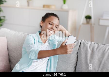 Fröhliches, jugendliches afro-amerikanisches Mädchen macht Selfie für soziale Netzwerke, sitzt zu Hause im Wohnzimmer auf dem Sofa Stockfoto