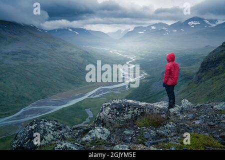 Weibliche Wanderin in roter Jacke mit Blick auf das abgelegene arktische Tal an einem bewölkten Tag im arktischen Sommer. Wandern im Sarek-Nationalpark, Schweden, mit dem Rapa-Fluss Stockfoto