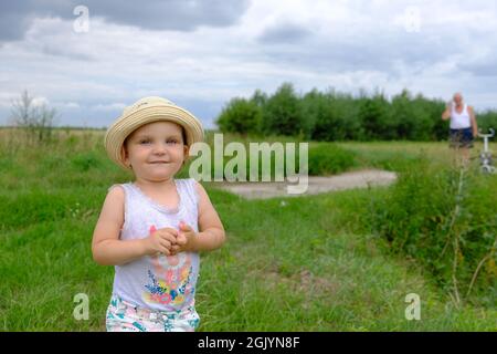 Porträt eines lächelnden kleinen Mädchens in einem Sommerhut, das auf einer grünen Wiese in Polen steht Stockfoto
