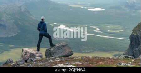 Weibliche Wanderin in blauer Jacke mit Blick auf das abgelegene arktische Tal an einem bewölkten Tag im arktischen Sommer. Wandern im Sarek-Nationalpark, Schweden, mit dem Rapa-Fluss Stockfoto