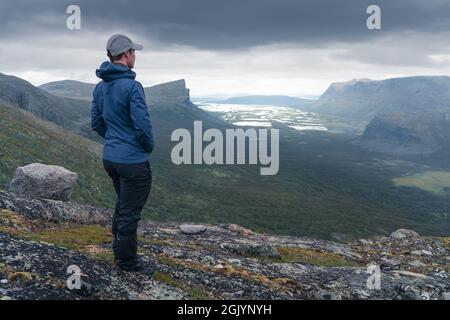 Weibliche Wanderin in blauer Jacke mit Blick auf das abgelegene arktische Tal an einem bewölkten Tag im arktischen Sommer. Wandern im Sarek-Nationalpark, Schweden, mit dem Rapa-Fluss Stockfoto