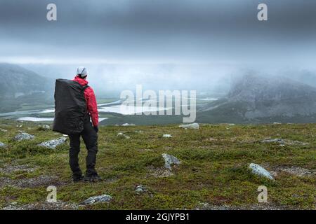 Weibliche Wanderin mit Blick auf das Rapa-Flusstal in der rauen arktischen Landschaft des Sarek-Nationalparks, Schweden, an einem sehr bewölkten und regnerischen Tag. Wandern in der Ferne Stockfoto