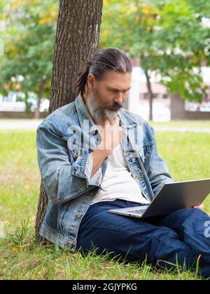 Im Stadtpark befindet sich ein Mann mittleren Alters mit einem schweren Bartbärtigen und einem Laptop. Er sitzt auf dem Gras im Park unter dem Baum. Vertikales Foto. Stockfoto