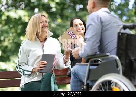 Behinderter Mann im Rollstuhl, der seinen Freundinnen im Park ein englisches Lehrbuch gab Stockfoto