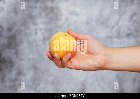 Die rechte Hand eines Mannes hält einen gelben Pfirsich auf einem strukturierten grauen Hintergrund. Stockfoto