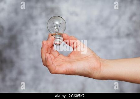 Die rechte Hand eines Mannes hält eine ausgelöschte Glühbirne aus Wolfram-Filament auf einem texturierten grauen Hintergrund. Stockfoto