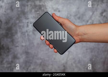 Die rechte Hand eines Mannes hält ein Smartphone mit ausgeschaltetem Bildschirm auf einem texturierten grauen Hintergrund. Stockfoto