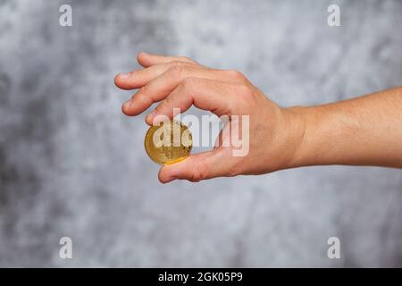 Die rechte Hand eines Mannes hält eine goldene Bitcoin-Münze auf einem strukturierten grauen Hintergrund. Stockfoto