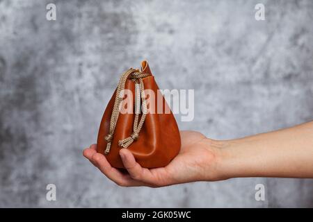 Die rechte Hand eines Mannes, der eine braune Ledertasche hält, die mit einer Schnur verschlossen ist, die Geld auf einem strukturierten grauen Hintergrund enthält. Stockfoto